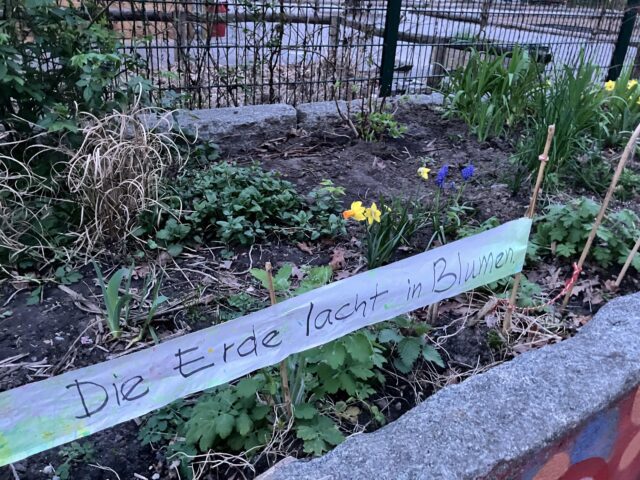 Ein handgemachtes Banner mit dem Text „Die Erde lacht Blumen“ auf den frisch bepflanzten Hochbeeten vor dem Spielhaus, Sesenheimer Straße.